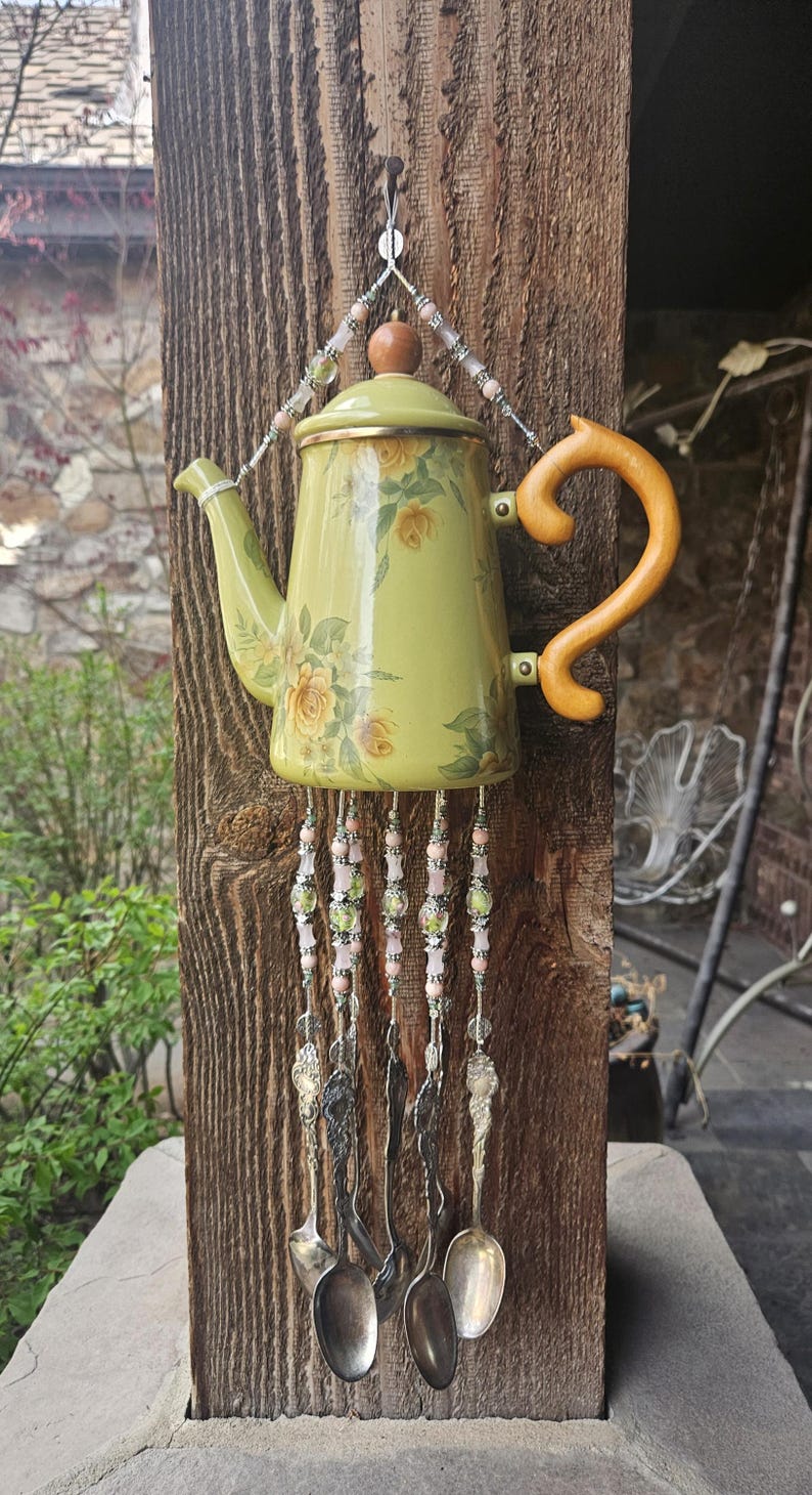 May include: A decorative wind chime crafted from a light green teapot featuring yellow floral patterns, a wooden handle, and a lid. Five silver-toned metal spoons hang from the base of the teapot, suspended by strings of pink and green beads.