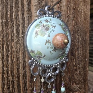 May include: A circular wall hanging featuring a light blue background with a floral pattern of purple violets and green leaves. A round, mottled brown knob is centered on the disc. Silver metal loops hang below, adorned with dangling beaded strands.