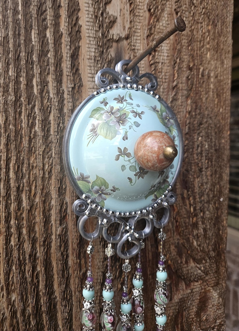 May include: A circular wall hanging featuring a light blue background with a floral pattern of purple violets and green leaves. A round, mottled brown knob is centered on the disc. Silver metal loops hang below, adorned with dangling beaded strands.