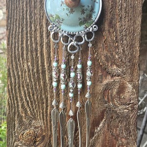 May include: A decorative wall hanging featuring a light blue ceramic lid with a floral pattern and a central wooden knob. Below the lid, five silver-toned metal spoons hang from silver wire strands adorned with light blue and purple beads.