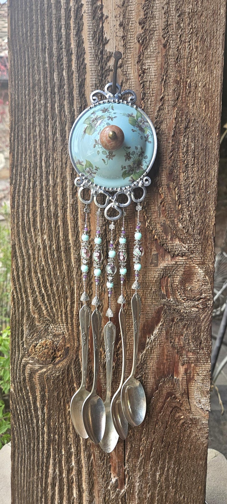 May include: A decorative wall hanging featuring a light blue ceramic lid with a floral pattern and a central wooden knob. Below the lid, five silver-toned metal spoons hang from silver wire strands adorned with light blue and purple beads.