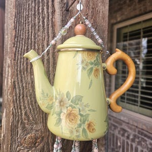 May include: A pale yellow enamel teapot wind chime featuring a floral rose pattern in shades of yellow and green. The teapot has a wooden handle and lid knob, suspended by a wire hanger adorned with pink and silver beads, with beaded strands.