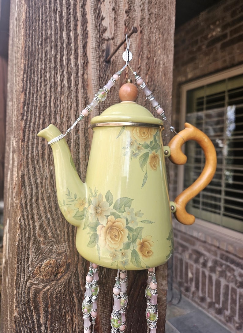 May include: A pale yellow enamel teapot wind chime featuring a floral rose pattern in shades of yellow and green. The teapot has a wooden handle and lid knob, suspended by a wire hanger adorned with pink and silver beads, with beaded strands.