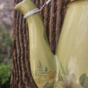 May include: A light olive green ceramic teapot featuring a floral pattern with yellow and green petals. A silver-toned metal chain adorned with small pink and green beads is attached to the spout, which has a metallic silver band at the top.