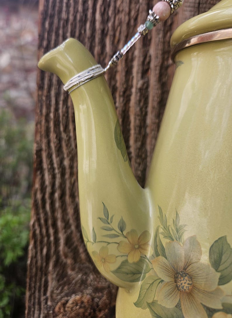 May include: A light olive green ceramic teapot featuring a floral pattern with yellow and green petals. A silver-toned metal chain adorned with small pink and green beads is attached to the spout, which has a metallic silver band at the top.