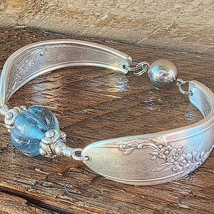 May include: A silver spoon bracelet with a blue glass bead and a silver ball charm. The bracelet is made from a vintage spoon and has a floral design.