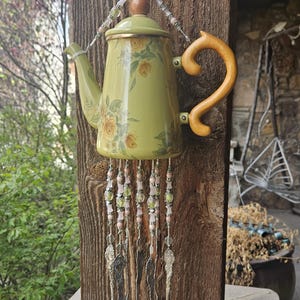 May include: A decorative wind chime crafted from a light green teapot with a yellow floral pattern, featuring a wooden handle and lid knob. Strands of pink and green beads hang from the base, ending in several silver-toned metal spoons.