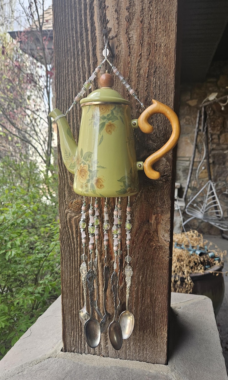 May include: A decorative wind chime crafted from a light green teapot with a yellow floral pattern, featuring a wooden handle and lid knob. Strands of pink and green beads hang from the base, ending in several silver-toned metal spoons.