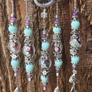 May include: A wind chime featuring five vertical strands suspended from silver metal loops. Each strand contains light blue matte beads, clear glass beads with pink and green floral patterns, small purple beads, and silver flower-shaped spacers, ending in silver spoon bowls.