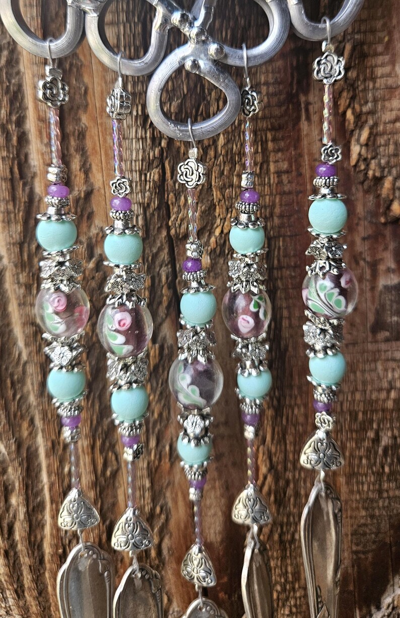 May include: A wind chime featuring five vertical strands suspended from silver metal loops. Each strand contains light blue matte beads, clear glass beads with pink and green floral patterns, small purple beads, and silver flower-shaped spacers, ending in silver spoon bowls.