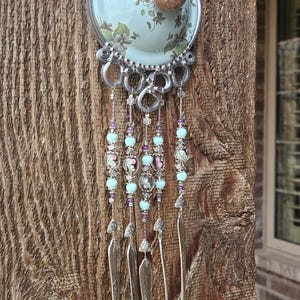 May include: A decorative wall hanging featuring a circular light blue ceramic plate with a floral pattern and a central round knob. Below the plate, four silver-toned metal spoons hang from silver wire strands adorned with light blue and purple beads.