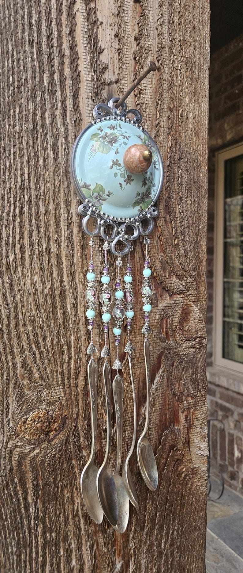 May include: A decorative wall hanging featuring a circular light blue ceramic plate with a floral pattern and a central round knob. Below the plate, four silver-toned metal spoons hang from silver wire strands adorned with light blue and purple beads.