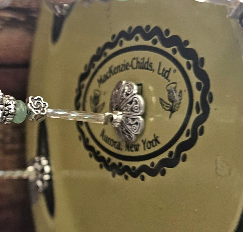 May include: A MacKenzie-Childs, Ltd. Aurora, New York branded object featuring a pale yellow circular base with a black scalloped border and thistle illustrations. A silver-toned metal decorative element with a floral motif is attached to the center.