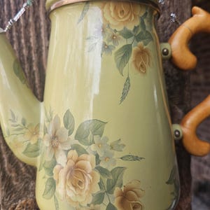 May include: A light green enamel coffee pot featuring a floral pattern of yellow roses and green leaves. The pot has a matching lid with a brown knob, a curved spout, and a light brown handle. Beaded decorative strands hang from the top and bottom.