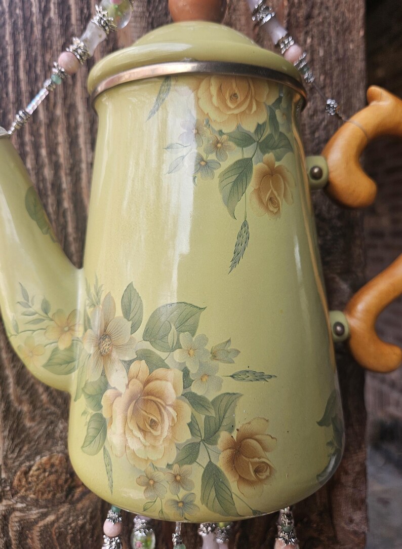 May include: A light green enamel coffee pot featuring a floral pattern of yellow roses and green leaves. The pot has a matching lid with a brown knob, a curved spout, and a light brown handle. Beaded decorative strands hang from the top and bottom.