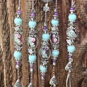 May include: A decorative hanging ornament featuring five vertical strands suspended from a silver-toned metal loop. Each strand is adorned with light blue matte beads, small purple beads, silver floral spacers, and clear glass beads with pink floral patterns, ending in silver metal spoon bowls.