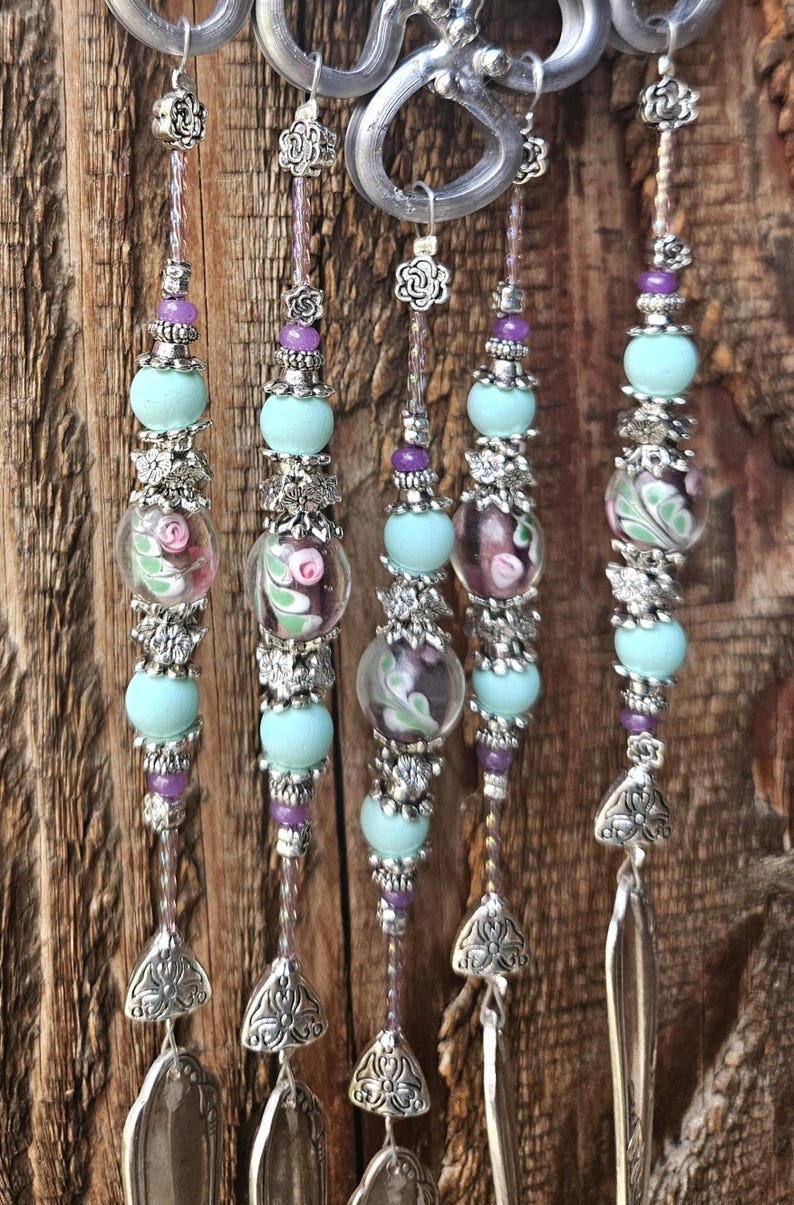 May include: A decorative hanging ornament featuring five vertical strands suspended from a silver-toned metal loop. Each strand is adorned with light blue matte beads, small purple beads, silver floral spacers, and clear glass beads with pink floral patterns, ending in silver metal spoon bowls.