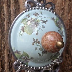 May include: A circular wall hook featuring a light blue dome decorated with purple violet flowers and green leaves. The center has a round, mottled orange-brown stone knob with a brass tip. The frame is silver metal with ornate scrollwork.