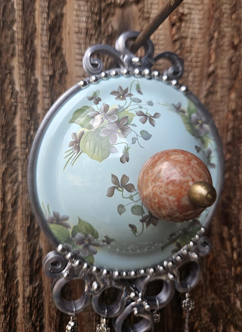May include: A circular wall hook featuring a light blue dome decorated with purple violet flowers and green leaves. The center has a round, mottled orange-brown stone knob with a brass tip. The frame is silver metal with ornate scrollwork.