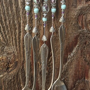 May include: A wind chime featuring five silver-toned metal spoons suspended from a decorative metal top. The spoons are connected by strands of light blue and floral-patterned glass beads, small silver spacers, and metal floral accents.
