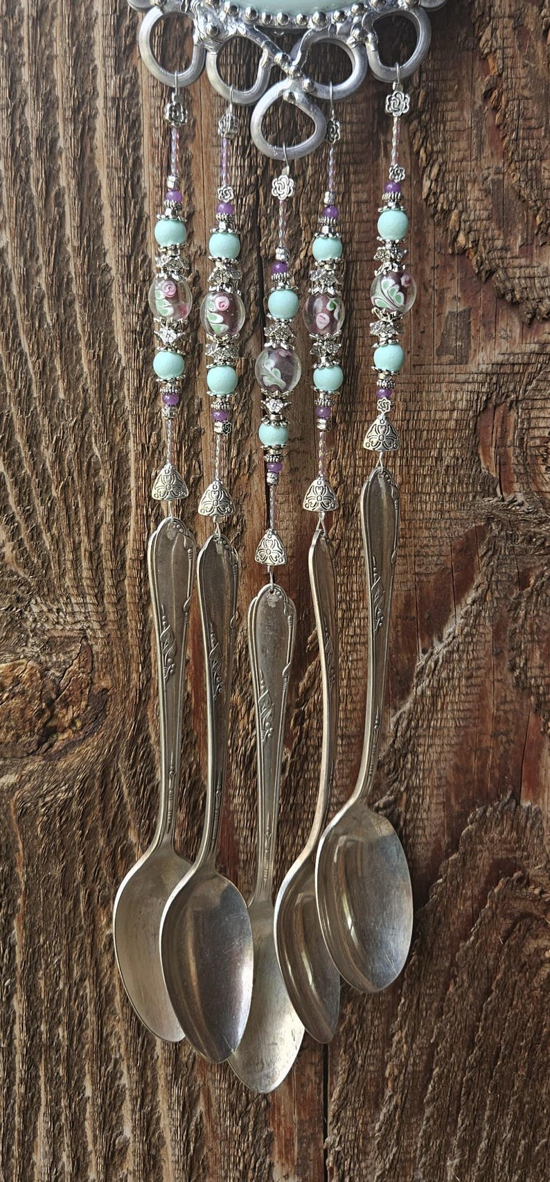 May include: A wind chime featuring five silver-toned metal spoons suspended from a decorative metal top. The spoons are connected by strands of light blue and floral-patterned glass beads, small silver spacers, and metal floral accents.
