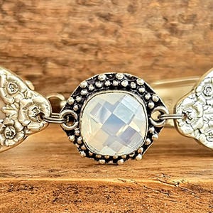 May include: A silver spoon bracelet with a large, clear, faceted crystal in the center. The spoon is decorated with floral designs.