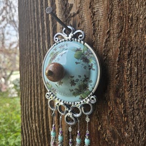 May include: A circular wall hanging featuring a light blue center with a floral pattern and a central brown knob. The piece is framed in silver metal with decorative scrollwork and hangs from a nail. Five beaded strands with light blue and purple accents hang from the bottom of the frame.