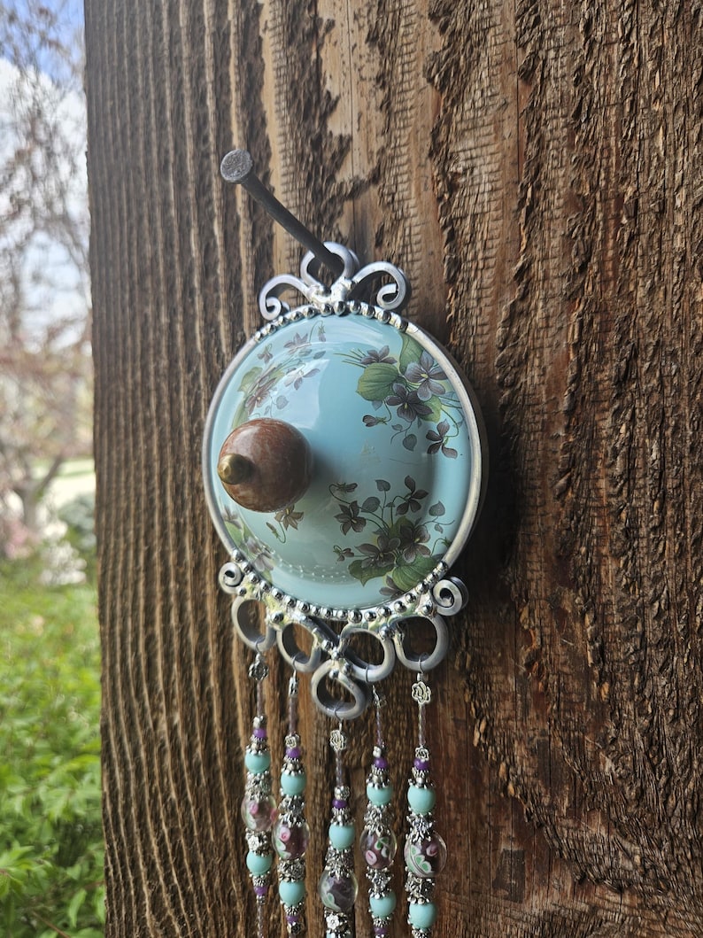May include: A circular wall hanging featuring a light blue center with a floral pattern and a central brown knob. The piece is framed in silver metal with decorative scrollwork and hangs from a nail. Five beaded strands with light blue and purple accents hang from the bottom of the frame.