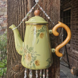 May include: A light green enamel teapot decorated with yellow rose floral patterns, featuring a wooden handle and a lid with a wooden knob. The teapot is suspended by a beaded wire hanger and adorned with dangling beaded strands at the base.