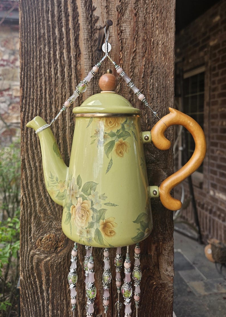 May include: A light green enamel teapot decorated with yellow rose floral patterns, featuring a wooden handle and a lid with a wooden knob. The teapot is suspended by a beaded wire hanger and adorned with dangling beaded strands at the base.