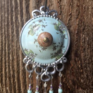 May include: A decorative wall hook featuring a circular light blue plate with a floral pattern, a central brown marble-like knob, and a silver-toned metal frame. Five dangling strands of light blue and clear beads with silver accents hang below.