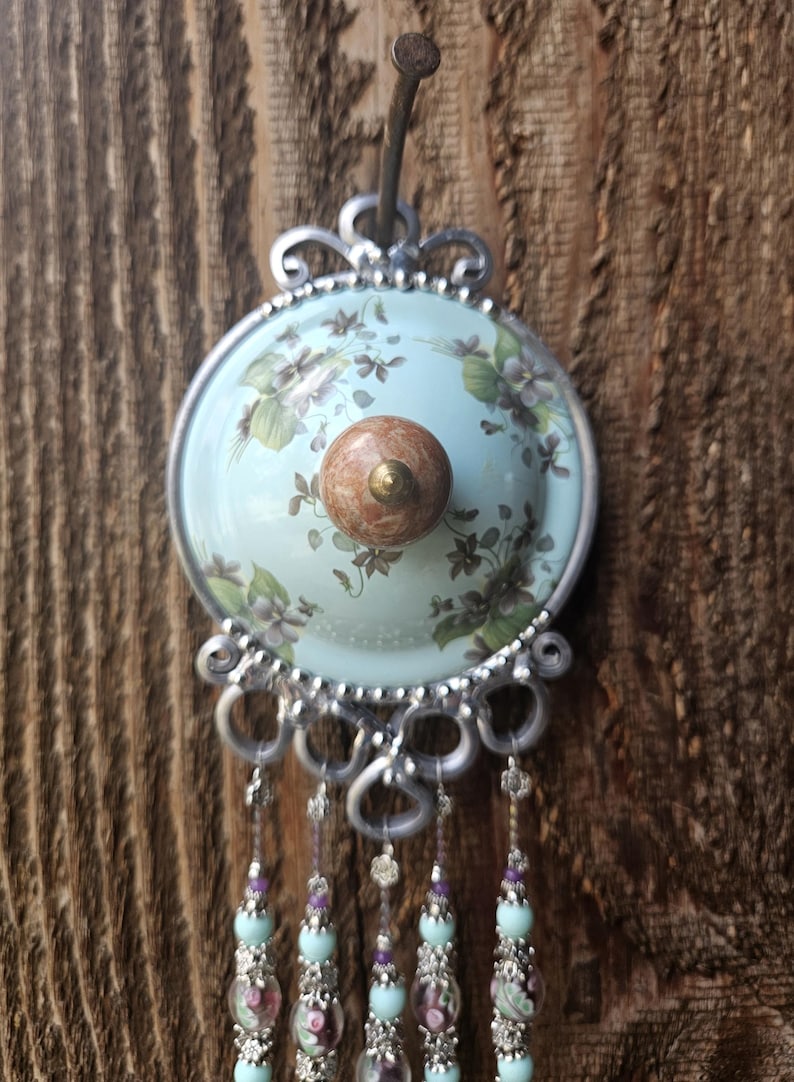 May include: A decorative wall hook featuring a circular light blue plate with a floral pattern, a central brown marble-like knob, and a silver-toned metal frame. Five dangling strands of light blue and clear beads with silver accents hang below.
