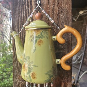 May include: A light green metal teapot featuring a yellow rose floral pattern, a wooden handle, and a wooden lid knob. The teapot is suspended by silver-toned wire adorned with pink and green beads, with additional beaded strands hanging below.