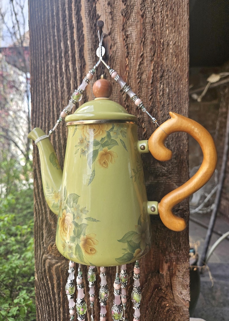 May include: A light green metal teapot featuring a yellow rose floral pattern, a wooden handle, and a wooden lid knob. The teapot is suspended by silver-toned wire adorned with pink and green beads, with additional beaded strands hanging below.