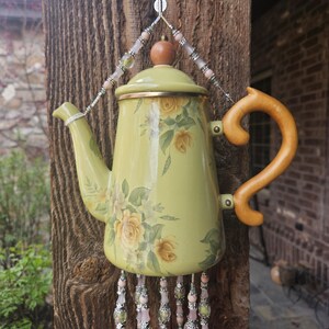 May include: A light green enamel teapot decorated with yellow roses, featuring a wooden handle and lid knob. The teapot is suspended by a wire adorned with pink and silver beads, with additional beaded strands hanging from the bottom base.