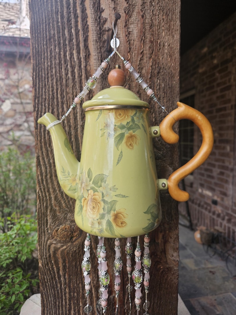 May include: A light green enamel teapot decorated with yellow roses, featuring a wooden handle and lid knob. The teapot is suspended by a wire adorned with pink and silver beads, with additional beaded strands hanging from the bottom base.
