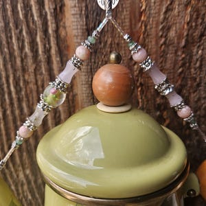 May include: A light green enamel teapot with a floral pattern, featuring a wooden knob on the lid. A decorative beaded handle attachment hangs above the lid, adorned with pink, clear, and green beads separated by silver-toned metal spacers.