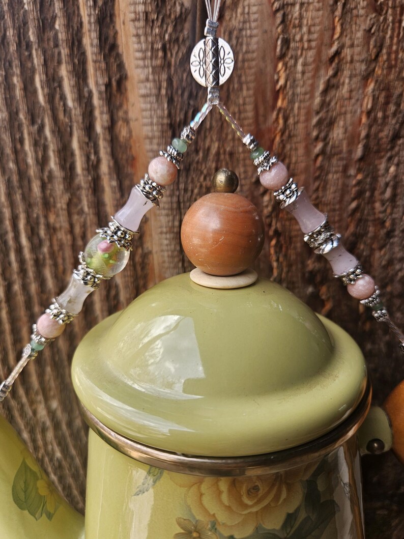 May include: A light green enamel teapot with a floral pattern, featuring a wooden knob on the lid. A decorative beaded handle attachment hangs above the lid, adorned with pink, clear, and green beads separated by silver-toned metal spacers.