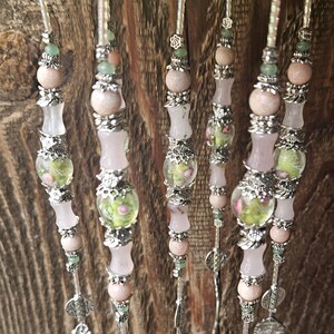 May include: A set of five vertical beaded strands featuring pale pink stone beads, translucent light green glass beads with floral patterns, and silver-toned metal spacers. Each strand is finished with a decorative silver-toned metal charm at the base.