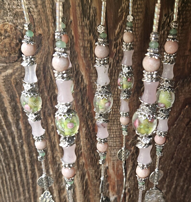May include: A set of five vertical beaded strands featuring pale pink stone beads, translucent light green glass beads with floral patterns, and silver-toned metal spacers. Each strand is finished with a decorative silver-toned metal charm at the base.