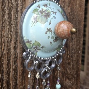 May include: A decorative wall hanging featuring a light blue circular plate with a floral pattern of purple violets and green leaves. A round, mottled brown stone knob sits in the center. Silver wire accents and dangling beaded strands hang below.