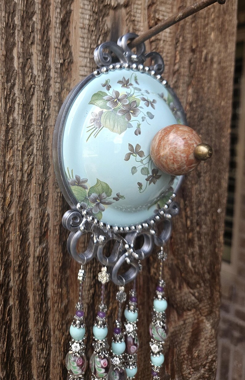 May include: A decorative wall hanging featuring a light blue circular plate with a floral pattern of purple violets and green leaves. A round, mottled brown stone knob sits in the center. Silver wire accents and dangling beaded strands hang below.