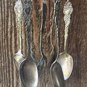 May include: A wind chime constructed from multiple vintage silver-toned spoons hanging vertically. Each spoon features an ornate, embossed handle with intricate floral and scrollwork patterns, suspended by thin metal chains and decorative circular metal beads.