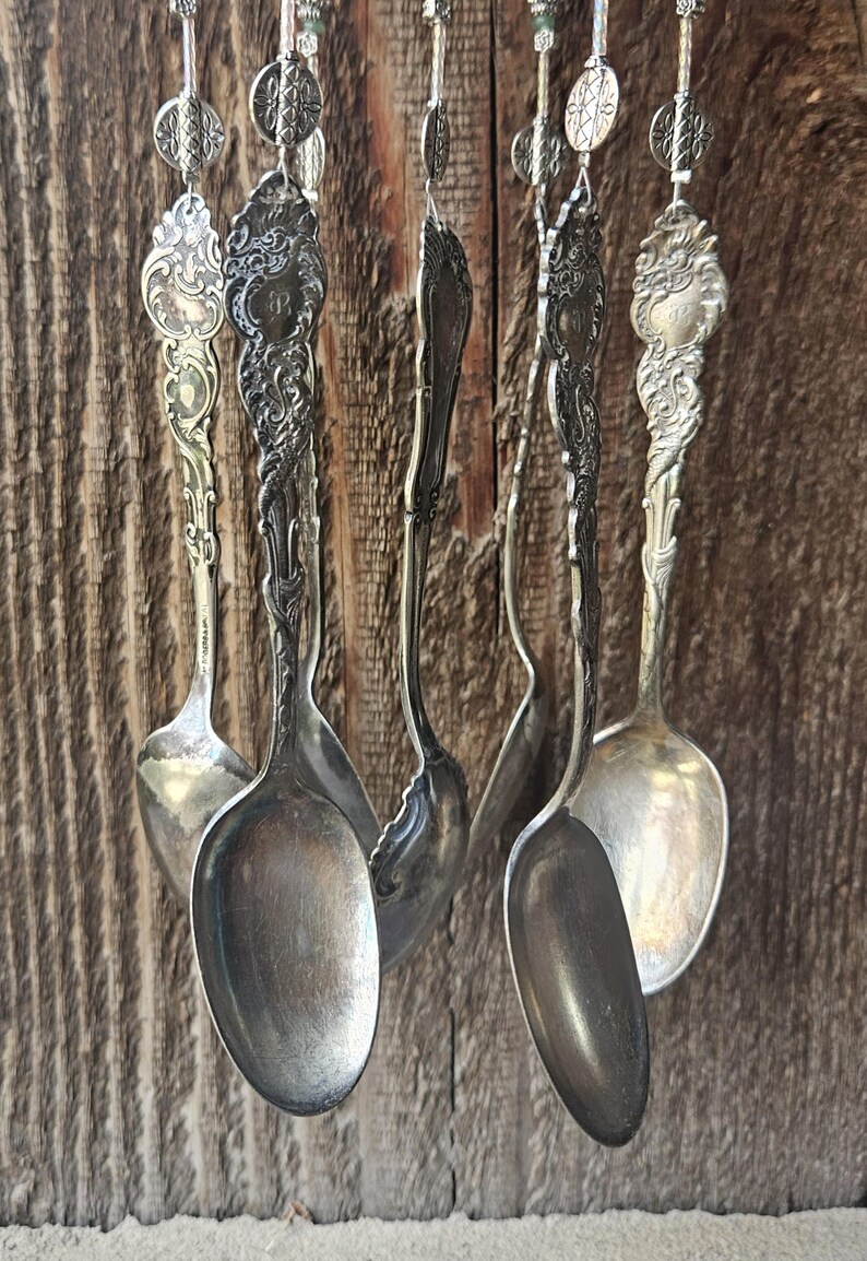May include: A wind chime constructed from multiple vintage silver-toned spoons hanging vertically. Each spoon features an ornate, embossed handle with intricate floral and scrollwork patterns, suspended by thin metal chains and decorative circular metal beads.