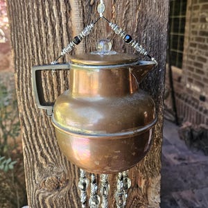May include: A copper-coloured metal teapot wind chime featuring a black handle and a clear glass lid knob. The teapot is suspended by a wire adorned with clear and black beads, with additional beaded strands hanging from the bottom of the vessel.