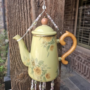 May include: A pale yellow teapot featuring a floral pattern with yellow roses and green leaves. The teapot has a wooden handle, a matching lid with a wooden knob, and is suspended by a wire hanger adorned with pink and green beads.