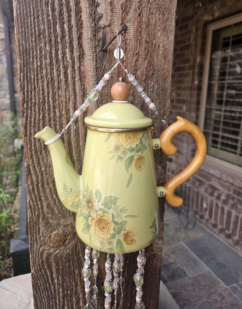 May include: A pale yellow teapot featuring a floral pattern with yellow roses and green leaves. The teapot has a wooden handle, a matching lid with a wooden knob, and is suspended by a wire hanger adorned with pink and green beads.