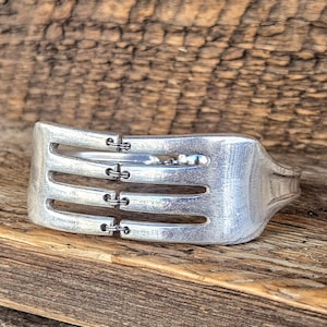 May include: A silver-colored fork bracelet with three horizontal slots and decorative accents. The bracelet is made from a repurposed fork and rests on a wooden surface, showcasing a rustic aesthetic.