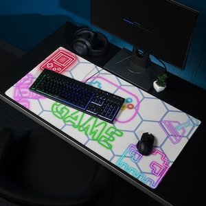 May include: A white gaming mouse pad with a black keyboard and mouse. The pad features a geometric pattern with neon-colored game-themed graphics, including a controller and the word "GAME".