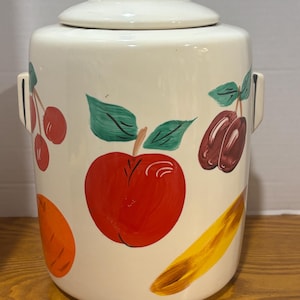 Könnte beinhalten: Cremefarbener Keramik-Keksdose mit Deckel und Griff. Die Dose ist mit handgemalten Früchten verziert, darunter ein roter Apfel, Kirschen, Pflaumen, eine Orangenscheibe und eine Banane. Die Dose hat zwei seitliche Griffe.